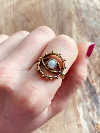 May include: A gold-toned ring with an eye motif, incorporating a central pale stone. The eye design is encircled by a ring with small, decorative beads. The ring is worn on a finger.